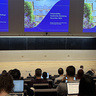 A university lecture hall with attendees seated at laptops facing a lecturer and three projected slides titled “Sustainability Leadership Workshop: December 2025.” A chalkboard behind the lecturer reads “Good Morning!”