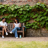 A man and a woman sit talking on a wooden bench in a garden area, with a brick wall covered in green vines behind them. A fern grows to the left of the bench, and a light blue cup sits on the gravel ground nearby.