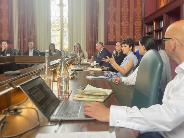 People sit around a large conference table in an ornate meeting room with red‑and‑gold wallpaper, tall windows, and bookshelves. Laptops, notebooks, and water bottles are on the table as the group participates in a formal discussion.