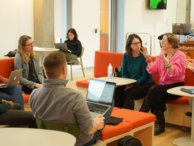 A group of seven people sit in a bright, modern workspace, gathered around small tables and orange benches with laptops open. One person gestures while speaking as others listen. In the background, another person works alone near a window and a reception-style desk is visible.