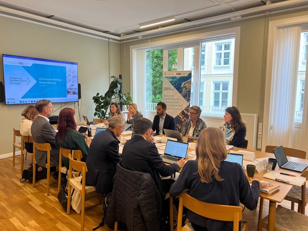 People sit around a wooden meeting table with laptops and papers during a formal working‑group session. A large screen displays “Corporate Leaders Group Europe – CLG Europe Working Group Meeting – May 2023,” and a standing banner beside it reads “Business Leadership for a Sustainable, Competitive and Resilient Europe.” The room is bright with large windows, wooden floors, and a plant in the corner.