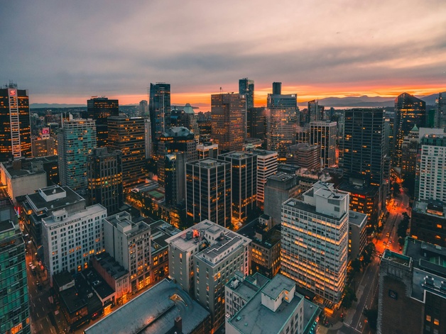 Aerial view of a modern city at sunset, with tall illuminated skyscrapers, dense urban streets below, and mountains in the distance under an orange‑pink sky.