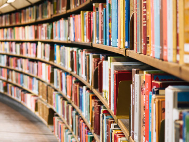 A large curved bookshelf filled with neatly arranged books in a spacious library. The shelves follow the curve of the room, with a metal railing and wooden floor visible, creating a sense of scale and organisation.