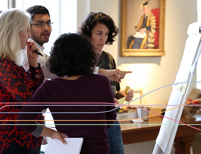 A group of four people stand around a flip chart in a bright meeting room. One person gestures toward the chart while another holds a marker and speaks, and the others watch with papers and sticky notes in hand. A framed portrait, a lamp, and a bowl of fruit sit in the background, giving the scene a collaborative workshop atmosphere.