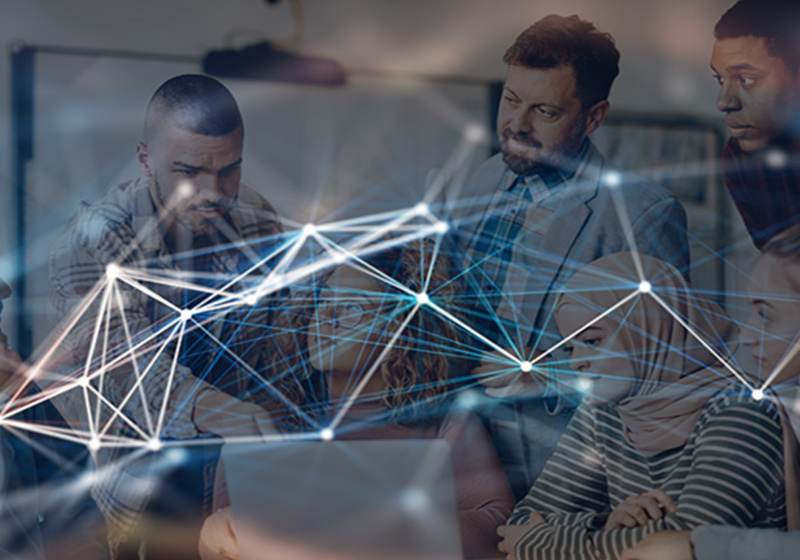 A group of six people gather closely around a laptop in a bright office setting, collaborating on a shared task. A digital network overlay of glowing lines and nodes appears across the image, symbolising data, connectivity, or AI‑driven teamwork.