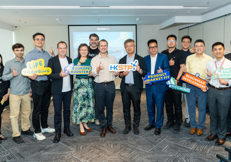 A group of thirteen people stand in a conference room giving thumbs‑up gestures and holding colourful signs with phrases such as “Life Changing,” “Europe Booster,” “Product Market Fit,” and “Pitching.” A presentation slide behind them reads “HKSTP Global Europe Booster.”