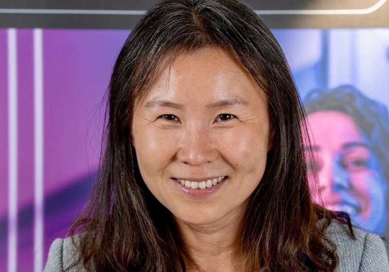Jie Zhou smiles at the camera in a grey blazer with the purple background from an innovation event