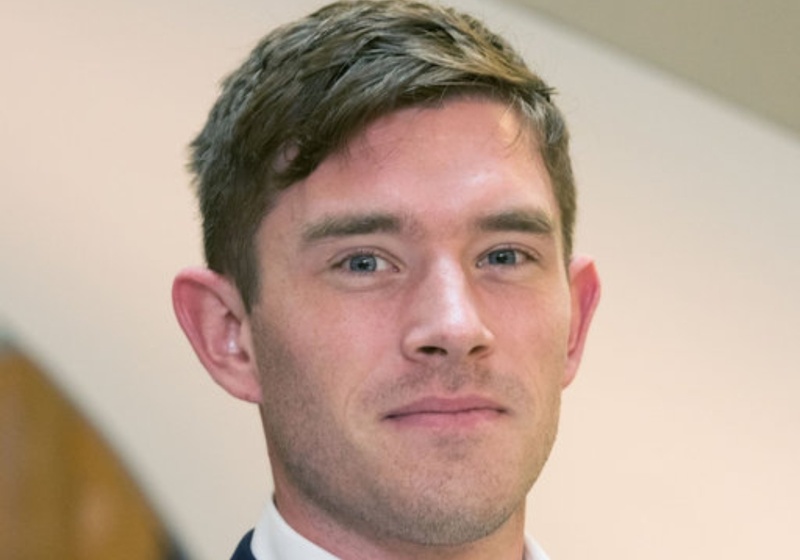 Dr Tom Norton smiles at the camera in a white shirt and navy blue blazer