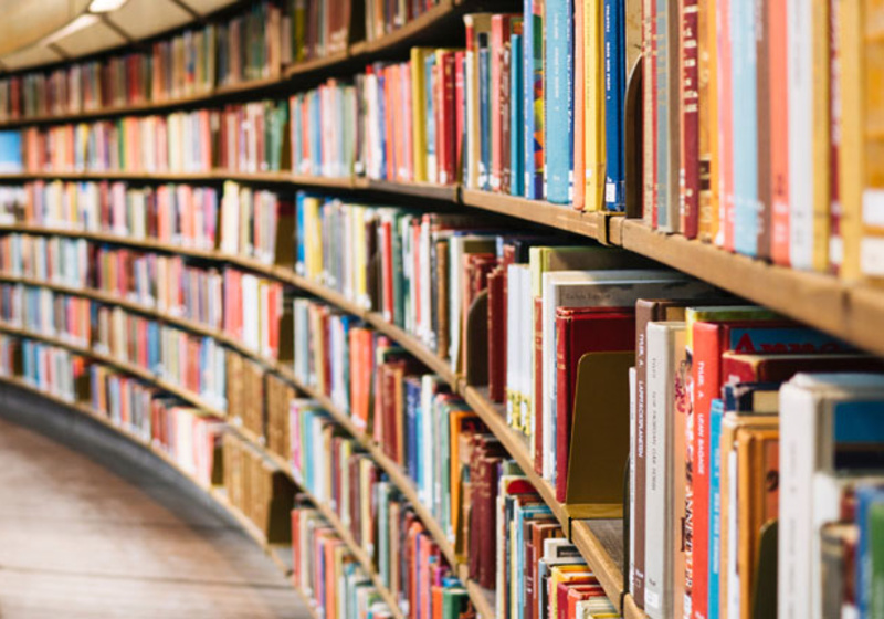 A large curved bookshelf filled with neatly arranged books in a spacious library. The shelves follow the curve of the room, with a metal railing and wooden floor visible, creating a sense of scale and organisation.