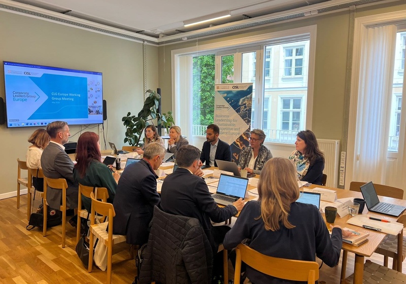 A group of people seated around a wooden meeting table with laptops and documents, watching a presentation slide titled “CLG Europe Working Group Meeting – May 2023” from the Cambridge Institute for Sustainability Leadership, with a banner behind them about business leadership for a sustainable and competitive Europe.