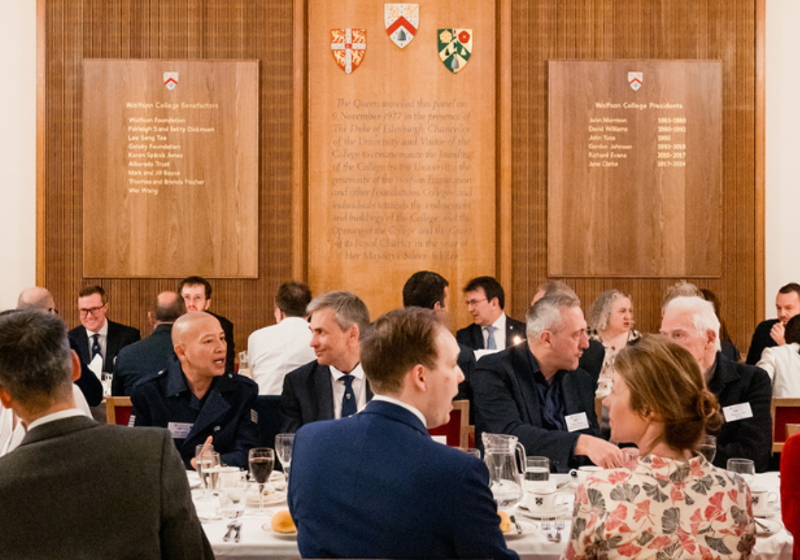 A formal dinner in a wood‑panelled hall at Wolfson College, with guests in suits and dresses seated at round tables beneath plaques listing benefactors and presidents, and three heraldic shields mounted above a central inscription.