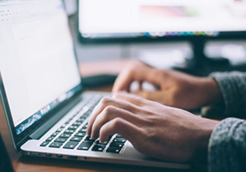Hands typing on a laptop keyboard at a desk, with a second monitor displaying text in the background.