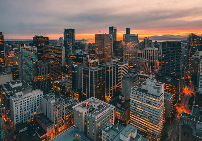 City skyline at sunset with tall buildings lit by warm streetlights and a glowing orange horizon.