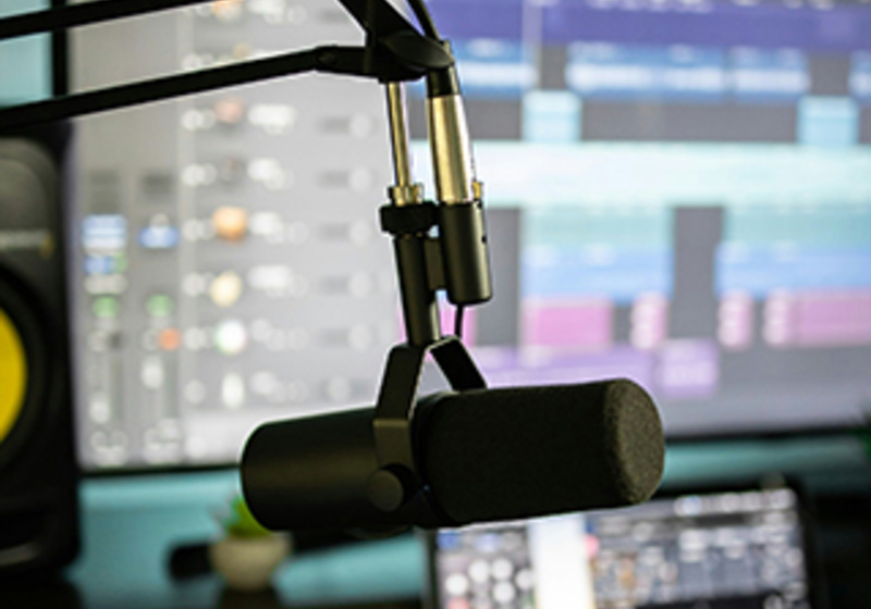 Close‑up of a podcast microphone on a boom arm with a computer screen showing audio editing software in the background.