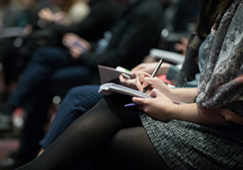 Person taking notes in a notebook during a seminar, seated among other attendees.