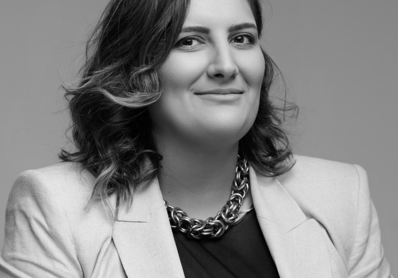 A black and white photo of Marija, in a smart blazer, dark top and thick necklace. Marija is smiling at the camera and looks professional. 
