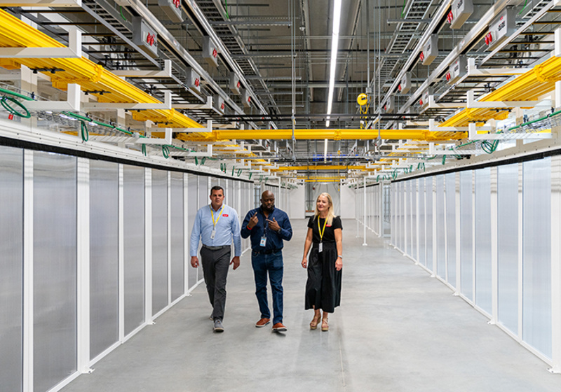 Three people walk through a brightly lit corridor at a Danfoss data centre facility, lined with rows of equipment enclosures. Overhead cable trays, power infrastructure and networking systems run the length of the facility, highlighting the scale and complexity of the digital infrastructure.