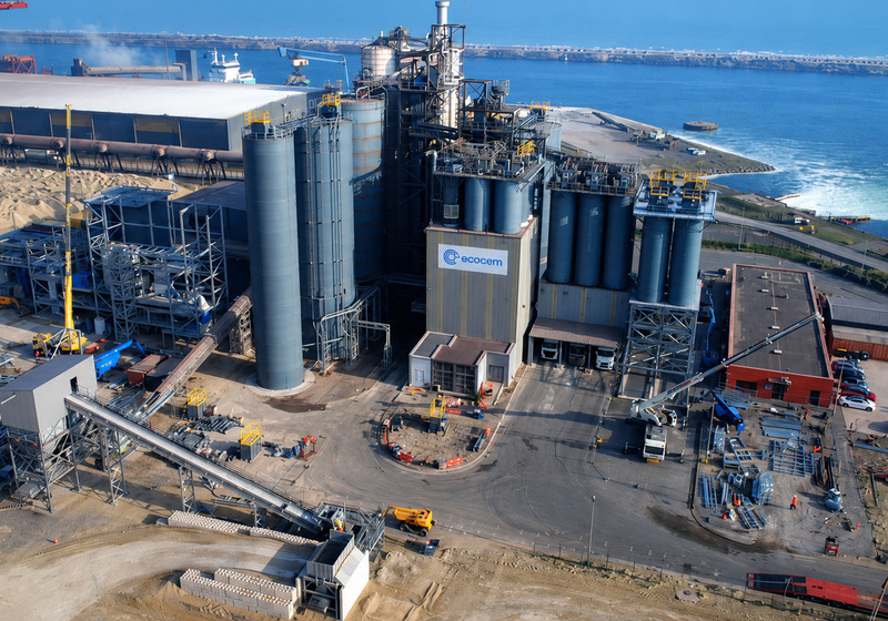 Aerial view of an industrial manufacturing facility located beside a coastal harbour. The site features large silos, processing equipment, conveyor systems, warehouses, and vehicles. Water, docks, and port infrastructure surround the facility, highlighting its connection to maritime transport and industrial operations.  Title text: Industrial manufacturing facil