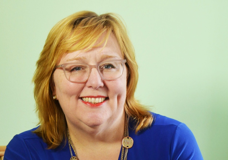 Jennifer, a white woman with light ginger hair, glasses, and a layered necklace is smiling at the camera in a blue dress.