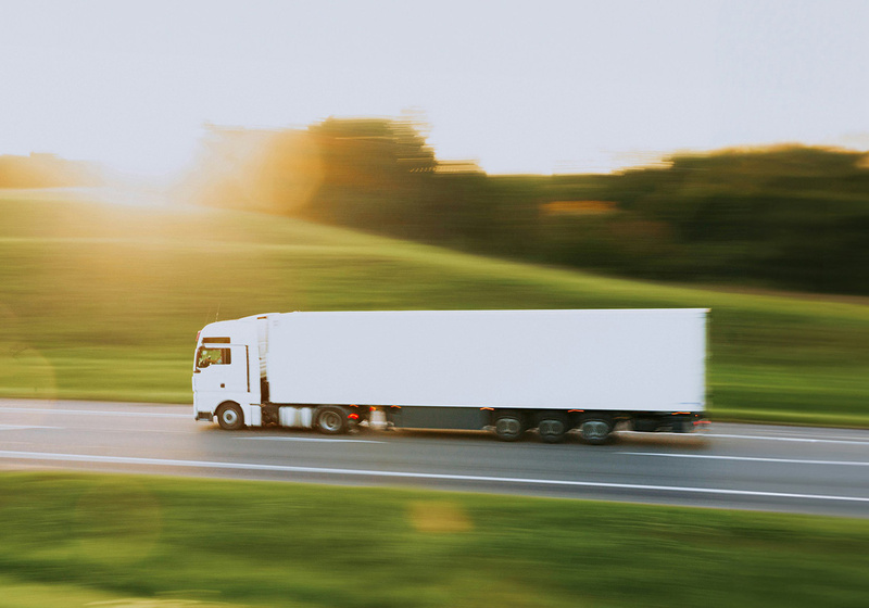 White articulated lorry travelling along a motorway through a rural landscape at sunset. Motion blur in the surrounding fields and trees emphasises the vehicle's movement, while warm sunlight illuminates the scene.