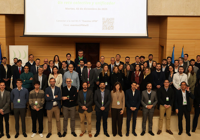 Group photo of approximately 70 conference attendees standing on a stage in a large wood-panelled auditorium beneath a projection screen displaying a Spanish-language presentation slide.