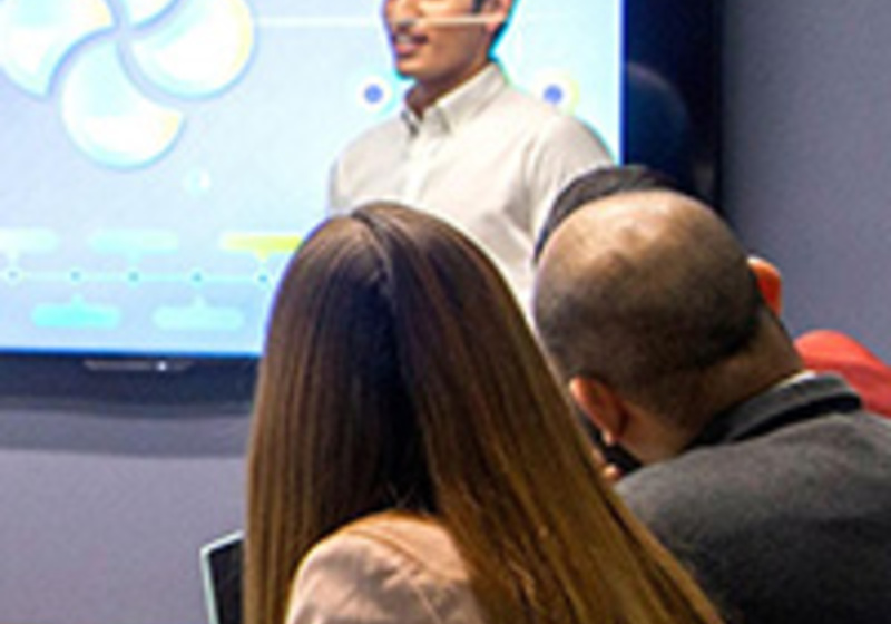 A presenter stands in front of a large screen displaying colourful charts and circular graphics, speaking to a group of people seated around a table in a meeting room.