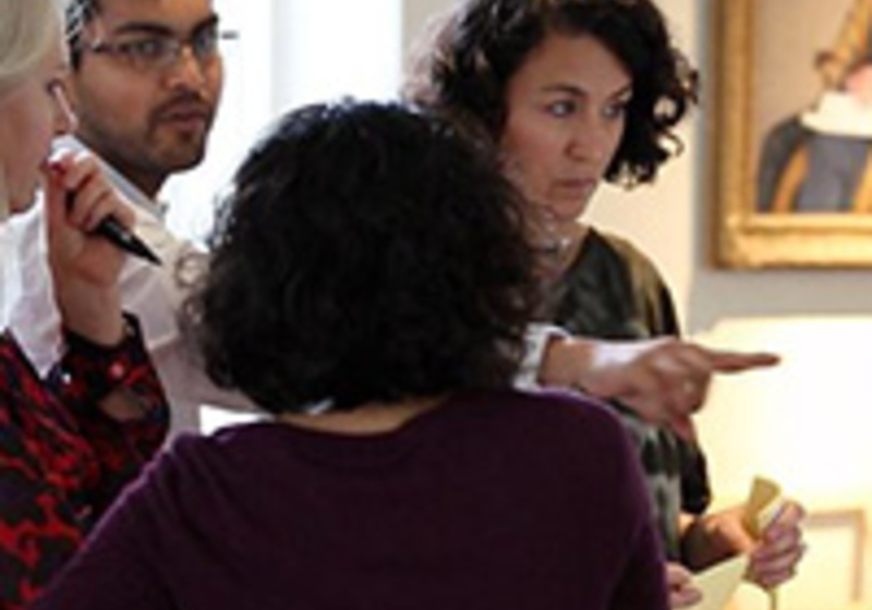 A group of four people stand together indoors, engaged in conversation. One person gestures toward something out of view while the others listen. Framed artwork hangs on the wall behind them, suggesting a professional or educational setting.