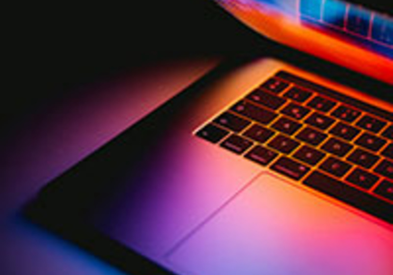 A partially open laptop is shown in close‑up, with vivid pink, purple, orange, and blue light reflecting across the keyboard and lower screen. The colourful glow highlights the sleek metallic surface and creates a modern, digital aesthetic.