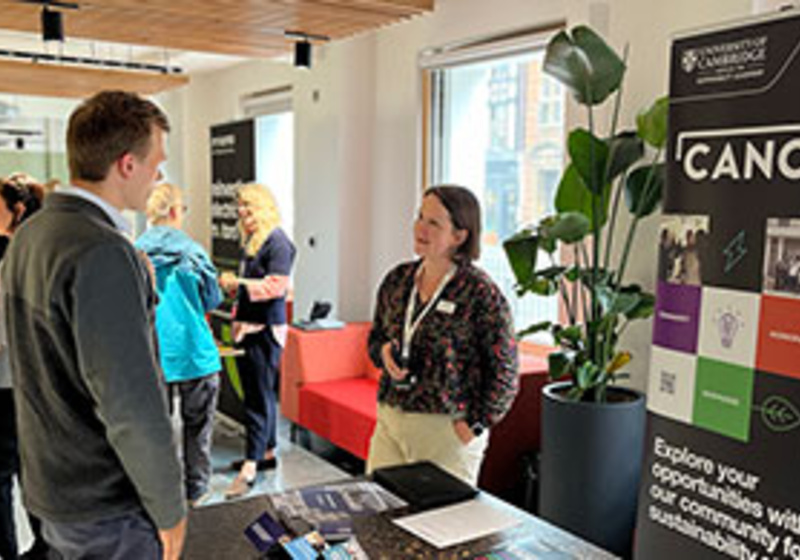 An indoor event space where two people are talking at a table beside a display stand featuring the University of Cambridge Canopy programme. Other attendees are gathered and conversing in the background.
