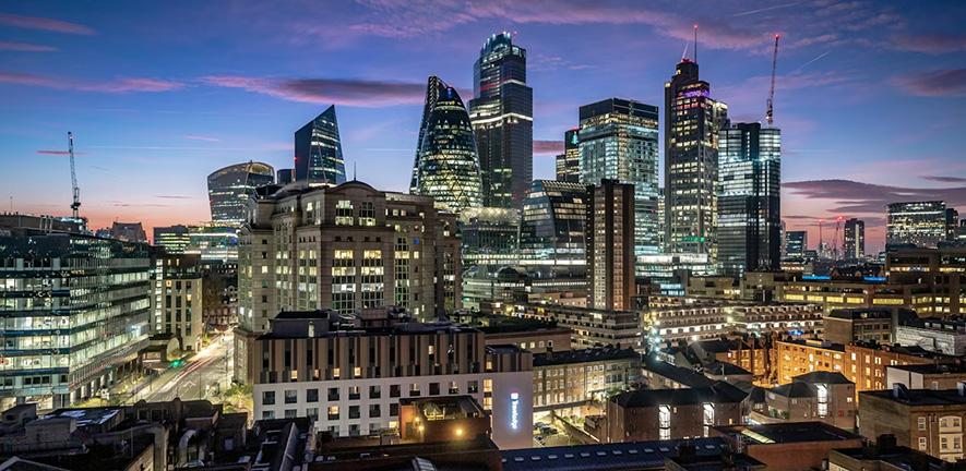 Twilight view of central London’s skyline, featuring The Gherkin, The Leadenhall Building, and other illuminated skyscrapers rising above older brick buildings under a blue‑purple dusk sky.