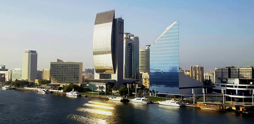 Modern waterfront skyline with several high‑rise buildings, including a curved glass tower and a triangular blue‑glass skyscraper, reflected in calm water with yachts moored in the foreground.