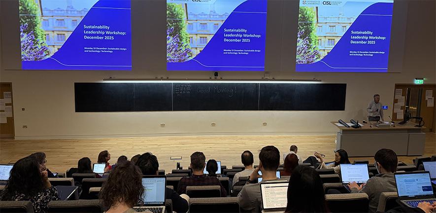 A university lecture hall with attendees seated at laptops facing a lecturer and three projected slides titled “Sustainability Leadership Workshop: December 2025.” A chalkboard behind the lecturer reads “Good Morning!”
