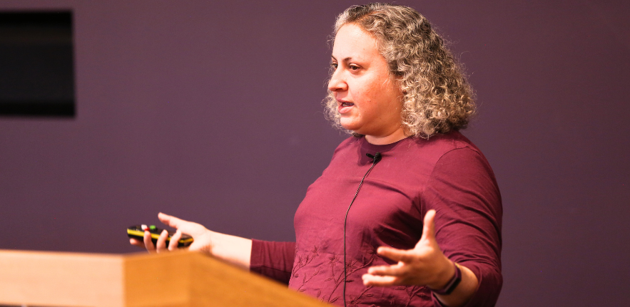 Dr Kayla Friedman A woman stood in front of a lecturn with hand outreached to her sides