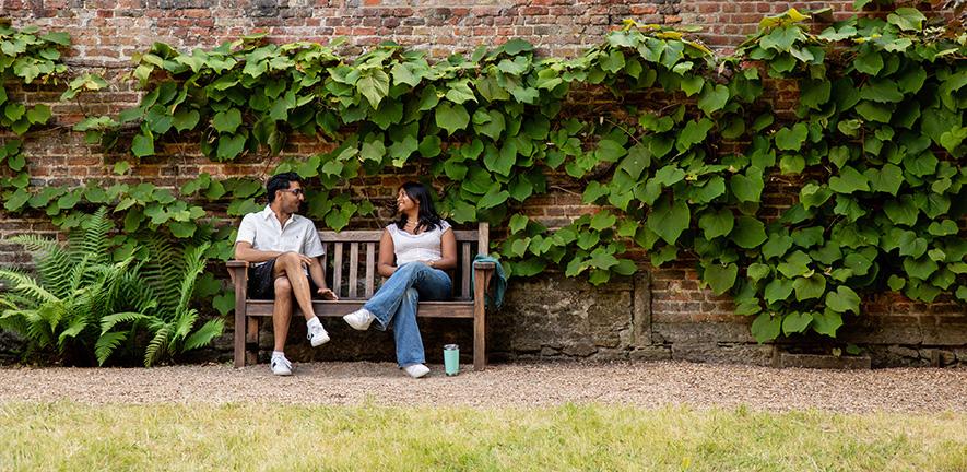A man and a woman sit talking on a wooden bench in a garden area, with a brick wall covered in green vines behind them. A fern grows to the left of the bench, and a light blue cup sits on the gravel ground nearby.