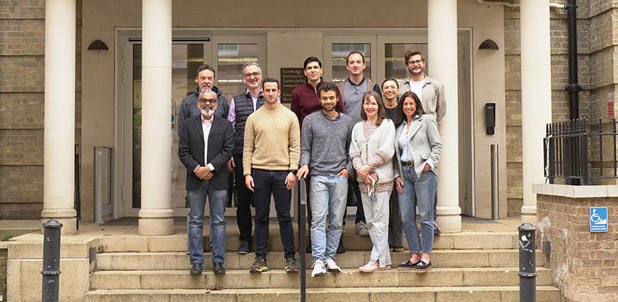 A group of people standing together on the steps outside a building with large pillars and glass doors. The group is arranged in two rows, with some standing on the upper step and others on the lower step. The building exterior features stone walls, white