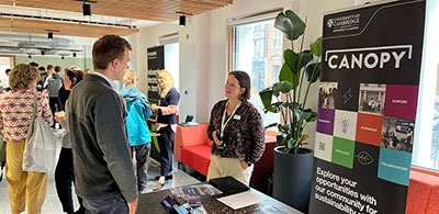 An indoor event space where two people are talking at a table beside a display stand featuring the University of Cambridge Canopy programme. Other attendees are gathered and conversing in the background.